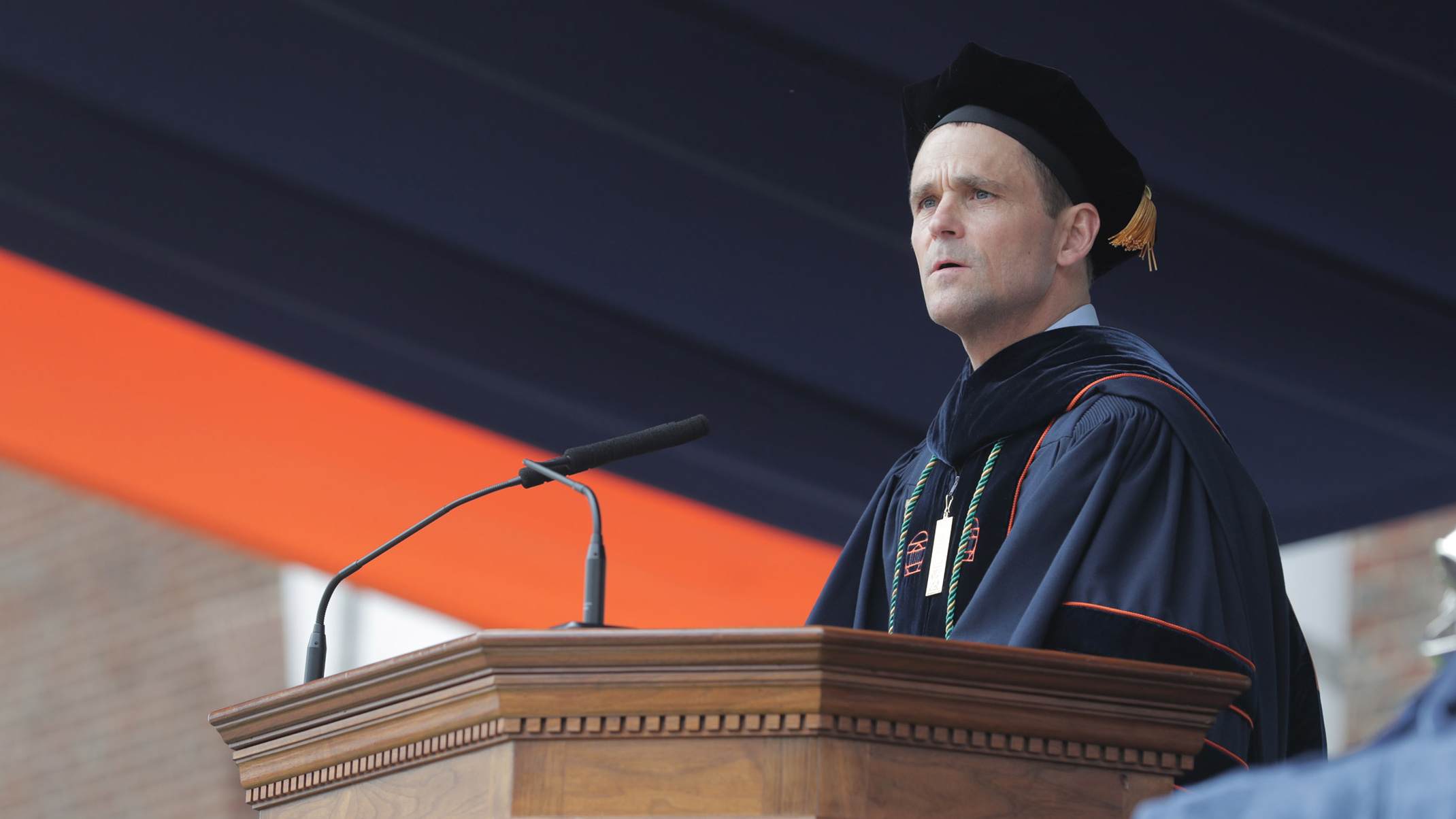 Jim Ryan speaking from a podium in Graduation attire outside